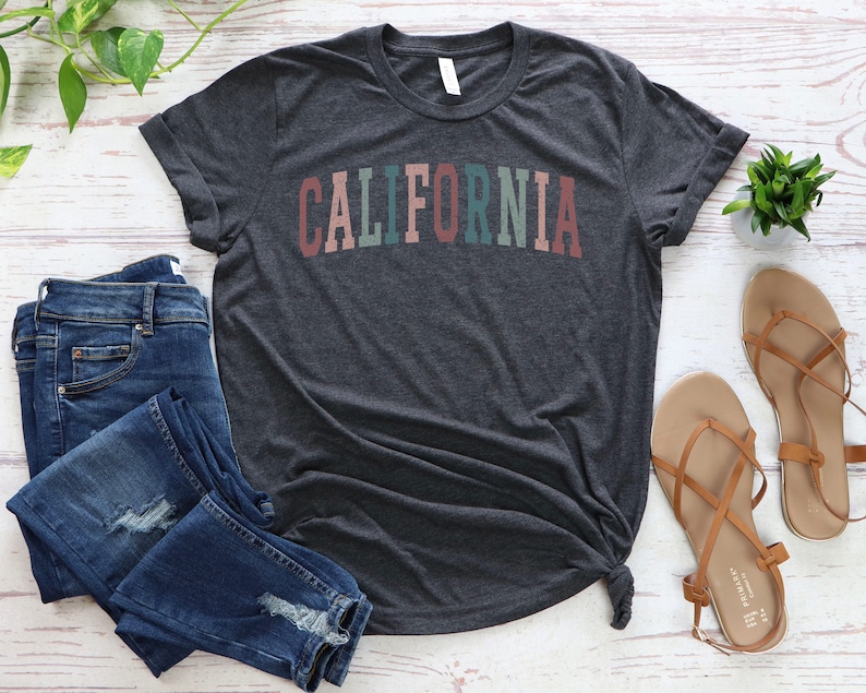 May include: A dark heather grey t-shirt with the word "CALIFORNIA" printed in rainbow colours across the front. The shirt is laid flat on a white wooden surface with a pair of blue jeans and brown sandals.