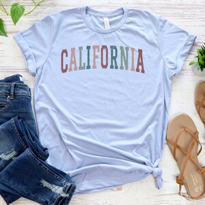 May include: A light blue t-shirt with the word "CALIFORNIA" printed in colourful letters. The shirt is laid out on a white wooden surface with a pair of brown sandals and blue jeans.