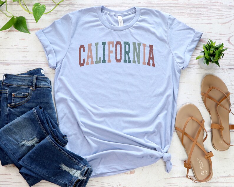 May include: A light blue t-shirt with the word "CALIFORNIA" printed in colourful letters. The shirt is laid out on a white wooden surface with a pair of brown sandals and blue jeans.