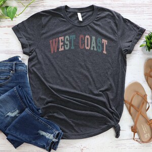 May include: A dark gray t-shirt with the text "WEST COAST" in colorful letters. The letters are in shades of pink, green, blue, and orange.