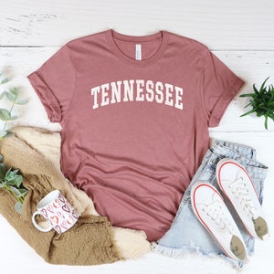 May include: A dusty rose colored t-shirt with the word "TENNESSEE" printed in white on the front. The shirt is laid flat on a white wooden surface with a pair of white sneakers, a mug, and a plant.