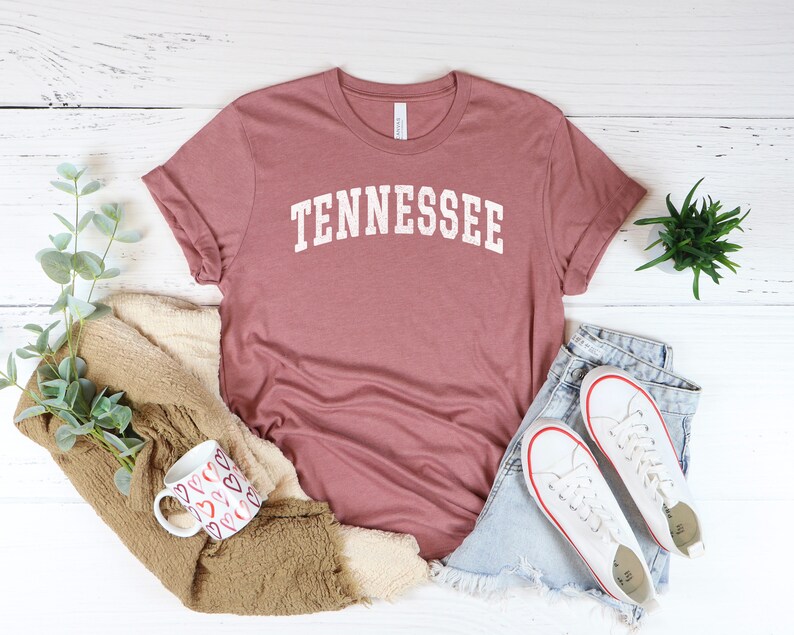 May include: A dusty rose colored t-shirt with the word "TENNESSEE" printed in white on the front. The shirt is laid flat on a white wooden surface with a pair of white sneakers, a mug, and a plant.