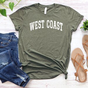 May include: A green t-shirt with the text "WEST COAST" in white lettering. The shirt is paired with blue jeans and brown sandals.