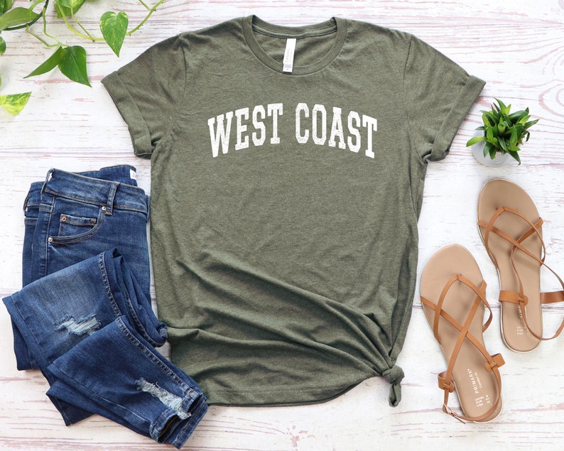 May include: A green t-shirt with the text "WEST COAST" in white lettering. The shirt is paired with blue jeans and brown sandals.