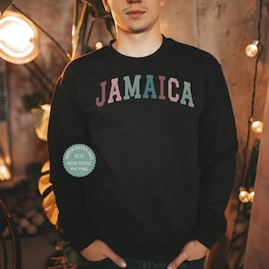 Puede incluir: Sudadera negra con la palabra "JAMAICA" impresa en un arco&iacute;ris a trav&eacute;s del pecho. La sudadera tiene una peque&ntilde;a etiqueta redonda verde en la manga izquierda que dice "Tintas a base de agua, Eco, No t&oacute;xico, Libre de PVC."