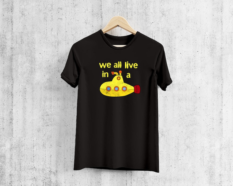 May include: Black t-shirt with the text "we all live in a" above a yellow submarine illustration. The submarine has blue portholes and a red tail. The shirt is hanging on a wooden hanger.