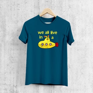 May include: Teal t-shirt with the text "we all live in a" above a yellow submarine graphic. The submarine has red and yellow accents. The shirt is hanging on a wooden hanger against a textured wall.