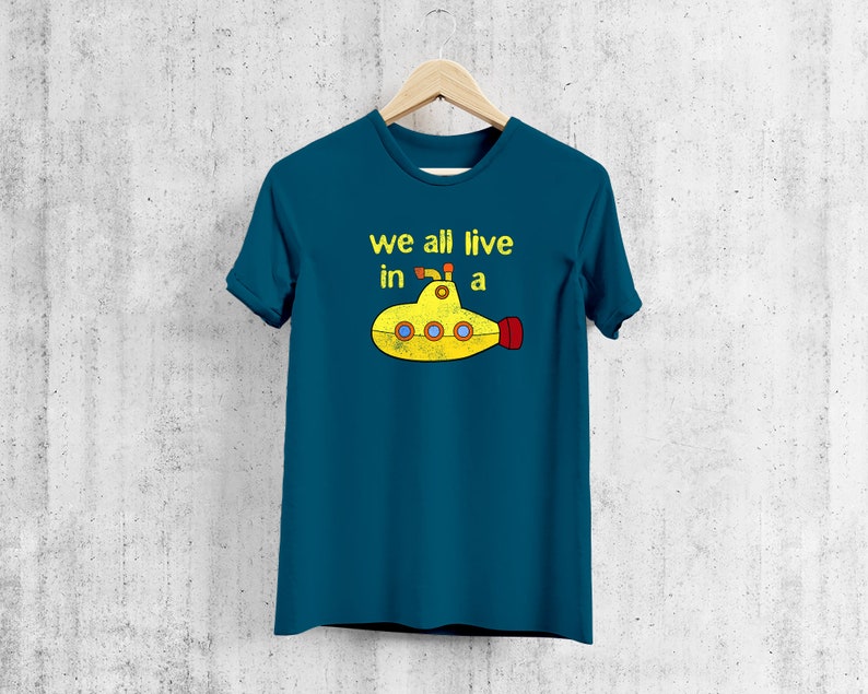 May include: Teal t-shirt with the text "we all live in a" above a yellow submarine graphic. The submarine has red and yellow accents. The shirt is hanging on a wooden hanger against a textured wall.