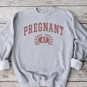 May include: Grey sweatshirt with the text "PREGNANT AF" in red lettering with a laurel wreath around the "AF".