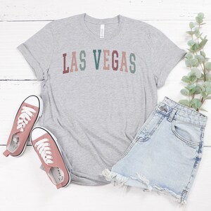 May include: A grey t-shirt with the text "LAS VEGAS" in a multi-coloured, arched design. The shirt is paired with light blue denim shorts and pink canvas trainers with white laces. A sprig of greenery is placed to the side.