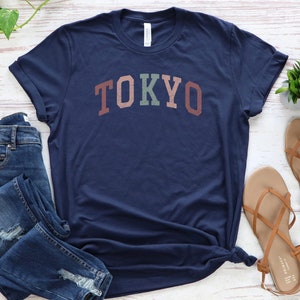 Pode incluir: Uma camiseta azul marinho com a palavra "TOKYO" impressa em arco-&iacute;ris na frente.