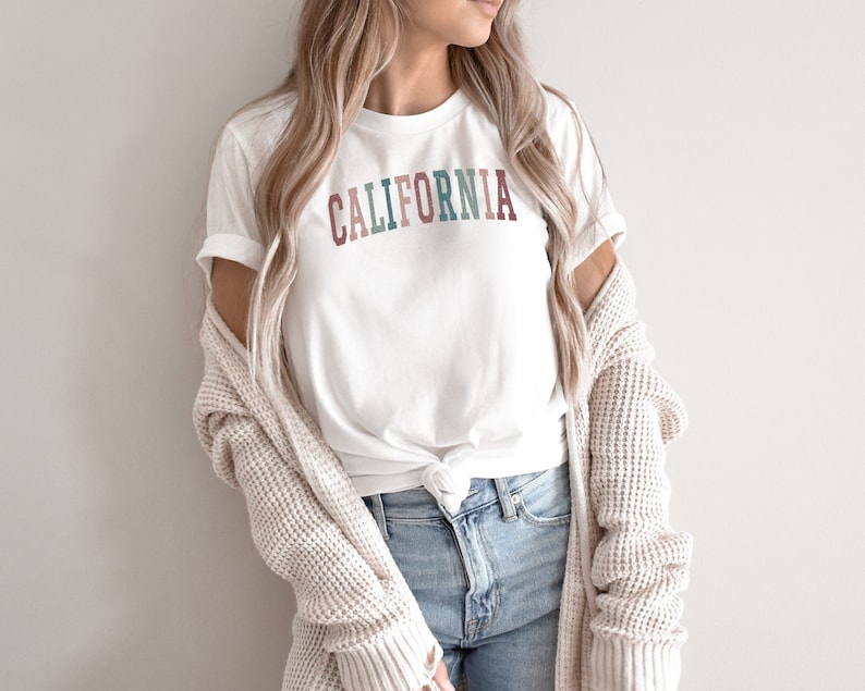 May include: A white t-shirt with the word "CALIFORNIA" printed in colourful letters. The letters are in shades of pink, blue, green, and brown.