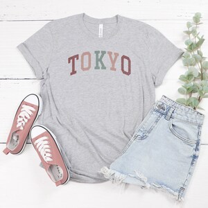 Pode incluir: Uma camiseta cinza com a palavra "TOKYO" impressa em letras coloridas. A camiseta &eacute; combinada com shorts jeans e t&ecirc;nis rosa.