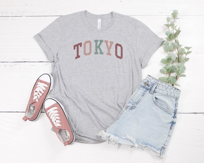 Pode incluir: Uma camiseta cinza com a palavra "TOKYO" impressa em letras coloridas. A camiseta &eacute; combinada com shorts jeans e t&ecirc;nis rosa.