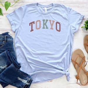 Pode incluir: Uma camiseta azul claro com a palavra "TOKYO" impressa em letras coloridas. A camiseta &eacute; combinada com jeans azul e sand&aacute;lias marrons.