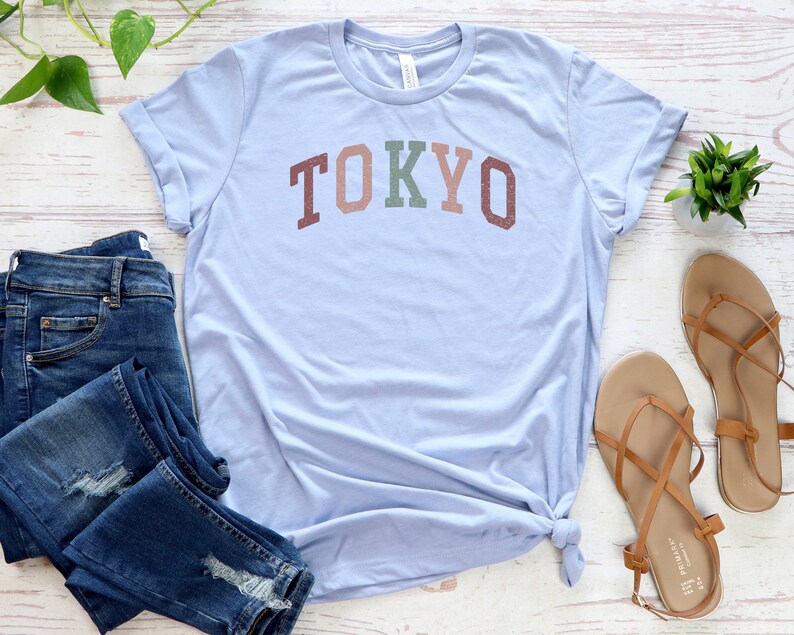 Pode incluir: Uma camiseta azul claro com a palavra "TOKYO" impressa em letras coloridas. A camiseta &eacute; combinada com jeans azul e sand&aacute;lias marrons.