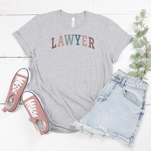 May include: A gray t-shirt with the word "LAWYER" printed in colorful letters. The shirt is paired with denim shorts and pink sneakers.