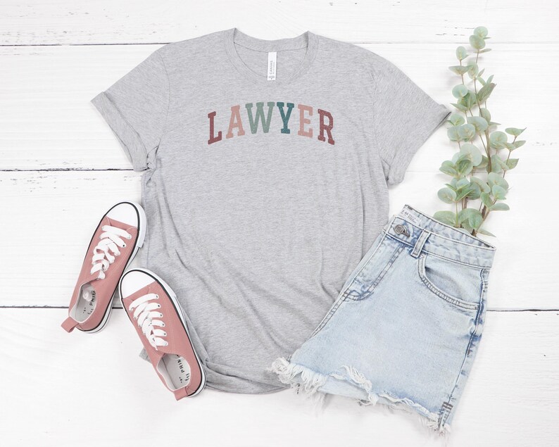 May include: A gray t-shirt with the word "LAWYER" printed in colorful letters. The shirt is paired with denim shorts and pink sneakers.
