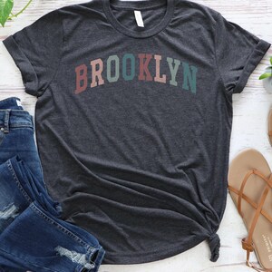 Op de afbeelding: Een donkergrijs T-shirt met een regenboogkleurige "BROOKLYN"-afbeelding.