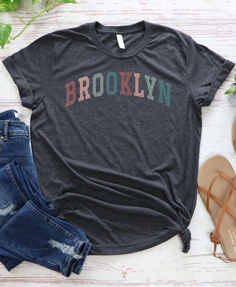 Op de afbeelding: Een donkergrijs T-shirt met een regenboogkleurige "BROOKLYN"-afbeelding.