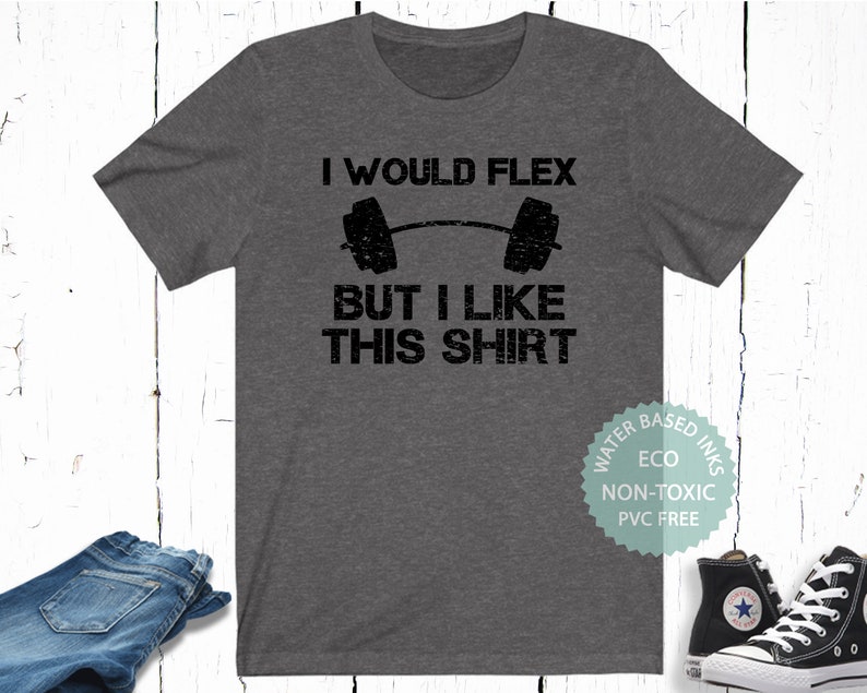 May include: A dark gray t-shirt with the text "I would flex but I like this shirt" and a black and white illustration of a barbell. The shirt is made with water-based, eco-friendly inks that are non-toxic and PVC free.