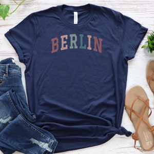 May include: A navy blue t-shirt with the word "BERLIN" printed across the chest in a multi-colored, arched design. The shirt is paired with blue jeans and brown sandals, arranged on a light wood-grain surface.