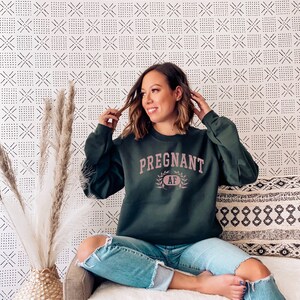 May include: A woman wearing a dark green sweatshirt with the text "PREGNANT AF" in pink lettering. She is sitting on a sofa with a white throw blanket and a patterned cushion.