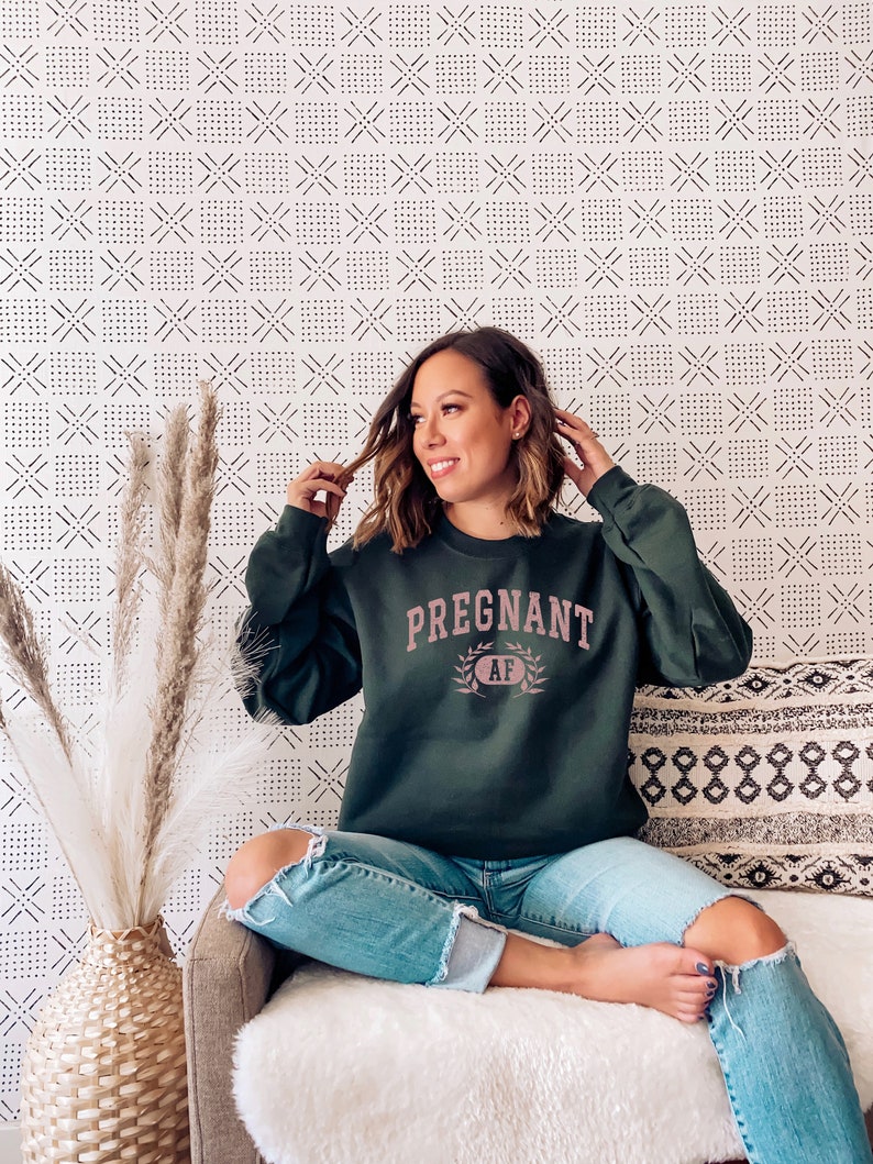 May include: A woman wearing a dark green sweatshirt with the text "PREGNANT AF" in pink lettering. She is sitting on a sofa with a white throw blanket and a patterned cushion.