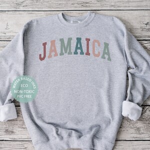 Puede incluir: Una sudadera gris con la palabra "JAMAICA" impresa en un arco&iacute;ris en la parte delantera. La sudadera est&aacute; hecha con tintas a base de agua, ecol&oacute;gicas, no t&oacute;xicas y sin PVC.