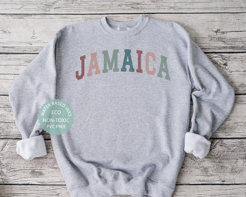 Puede incluir: Una sudadera gris con la palabra "JAMAICA" impresa en un arco&iacute;ris en la parte delantera. La sudadera est&aacute; hecha con tintas a base de agua, ecol&oacute;gicas, no t&oacute;xicas y sin PVC.