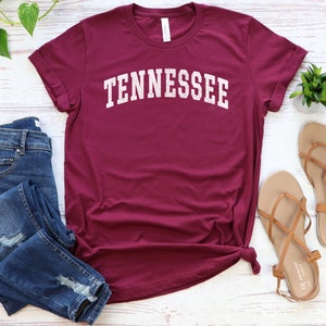 May include: A maroon t-shirt with the word "TENNESSEE" in white text on the front. The shirt is laid flat on a white wooden surface with a pair of brown sandals and blue jeans.