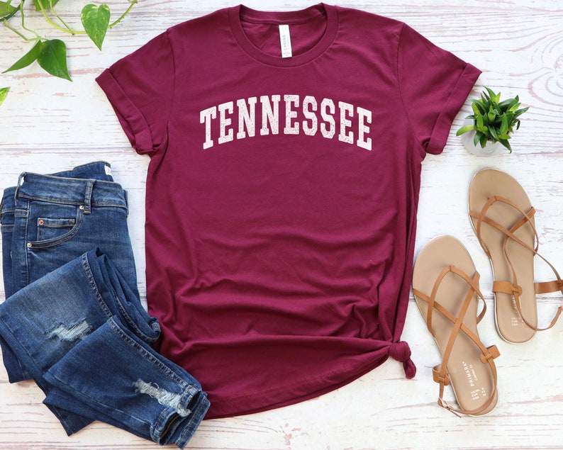 May include: A maroon t-shirt with the word "TENNESSEE" in white text on the front. The shirt is laid flat on a white wooden surface with a pair of brown sandals and blue jeans.
