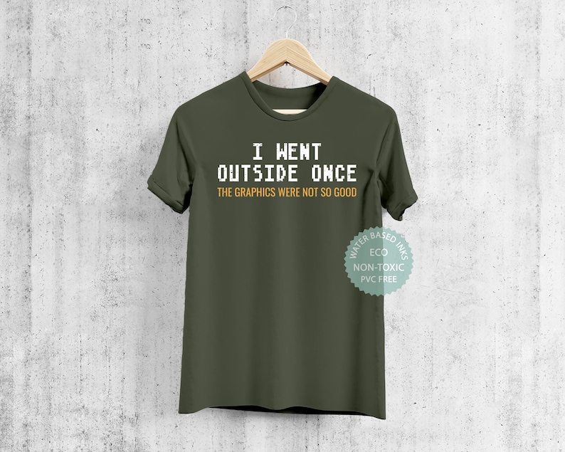 May include: A green t-shirt with the text "I WENT OUTSIDE ONCE THE GRAPHICS WERE NOT SO GOOD" in white pixelated font. The text is centered on the shirt. The shirt has a white tag on the bottom right corner that says "WATER BASED INKS ECO NON-TOXIC PVC FREE".
