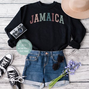 Puede incluir: Sudadera negra con la palabra "JAMAICA" en colores arco&iacute;ris. La sudadera est&aacute; colocada sobre un fondo de madera blanca con un par de zapatillas negras, pantalones cortos de mezclilla azul, un sombrero marr&oacute;n, una c&aacute;mara y un ramo de flores moradas.