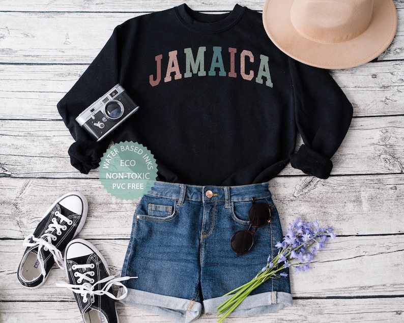 Puede incluir: Sudadera negra con la palabra "JAMAICA" en colores arco&iacute;ris. La sudadera est&aacute; colocada sobre un fondo de madera blanca con un par de zapatillas negras, pantalones cortos de mezclilla azul, un sombrero marr&oacute;n, una c&aacute;mara y un ramo de flores moradas.