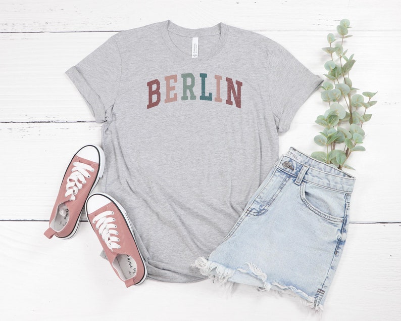 May include: A heather gray t-shirt with the word "BERLIN" in a multi-colored arched design. The shirt is paired with light blue denim shorts and pink canvas sneakers. A sprig of greenery is also included in the image.