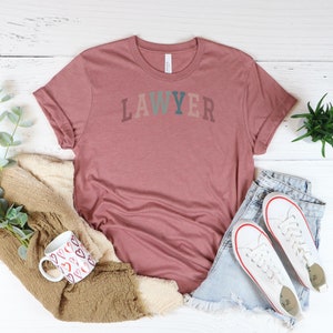 May include: A mauve t-shirt with the word "LAWYER" printed in colorful letters. The shirt is laid out on a white wooden surface with a mug, a blanket, and a pair of jeans.