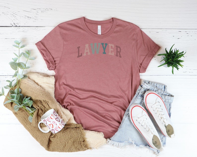 May include: A mauve t-shirt with the word "LAWYER" printed in colorful letters. The shirt is laid out on a white wooden surface with a mug, a blanket, and a pair of jeans.