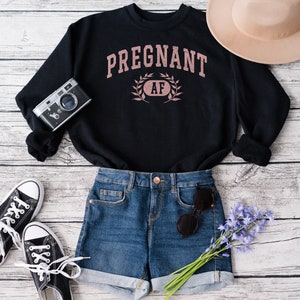 May include: Black sweatshirt with the text "PREGNANT AF" in pink lettering with a wreath around the "AF". The sweatshirt is laid out on a white wood background with a pair of black trainers, a pair of blue denim shorts, a brown hat, and a vintage camera.