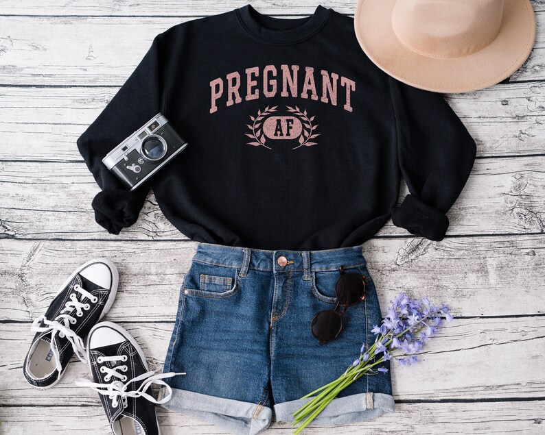 May include: Black sweatshirt with the text "PREGNANT AF" in pink lettering with a wreath around the "AF". The sweatshirt is laid out on a white wood background with a pair of black trainers, a pair of blue denim shorts, a brown hat, and a vintage camera.