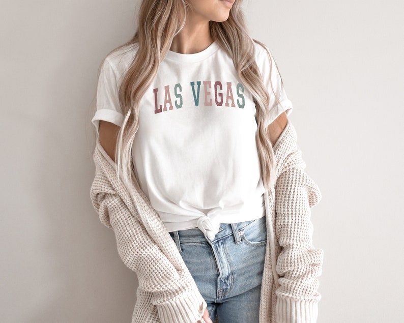 May include: White t-shirt with the text "LAS VEGAS" in colourful letters. The shirt is paired with blue jeans and a cream-coloured cardigan. The overall style is casual and relaxed.