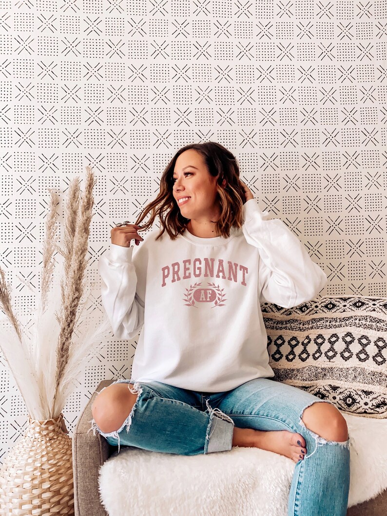 May include: A white sweatshirt with the text "PREGNANT AF" in pink lettering with a laurel wreath around the text. The sweatshirt is being worn by a person sitting on a sofa.