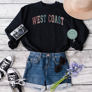 Puede incluir: Una sudadera negra con el texto "WEST COAST" en letras de colores. La sudadera est&aacute; colocada sobre una superficie de madera blanca con un par de zapatillas negras, unos pantalones cortos de mezclilla azul, un sombrero marr&oacute;n, una c&aacute;mara y un ramo de flores moradas.