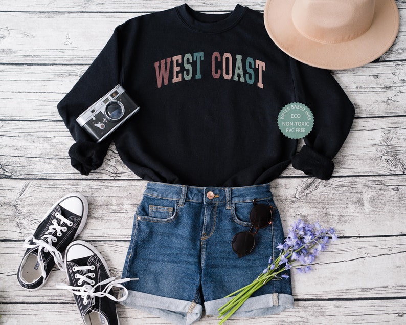 Puede incluir: Una sudadera negra con el texto "WEST COAST" en letras de colores. La sudadera est&aacute; colocada sobre una superficie de madera blanca con un par de zapatillas negras, unos pantalones cortos de mezclilla azul, un sombrero marr&oacute;n, una c&aacute;mara y un ramo de flores moradas.