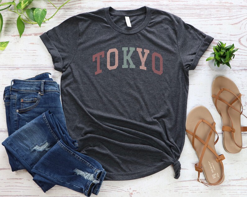 Pode incluir: Uma camiseta cinza escura mesclada com a palavra "TOKYO" impressa em letras coloridas. A camiseta est&aacute; colocada sobre uma superf&iacute;cie de madeira branca com um par de cal&ccedil;as de ganga azuis e sand&aacute;lias marrons.