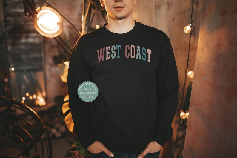 Puede incluir: Una sudadera negra con el texto "WEST COAST" impreso en un degradado arco&iacute;ris en el pecho. La sudadera est&aacute; hecha con tintas a base de agua que son ecol&oacute;gicas, no t&oacute;xicas y sin PVC.