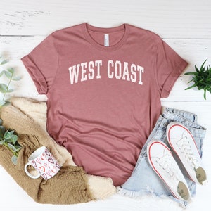 May include: A mauve colored t-shirt with the text "WEST COAST" printed in white on the front. The t-shirt is laid flat on a white wooden surface with a pair of white sneakers, a mug, and a plant.