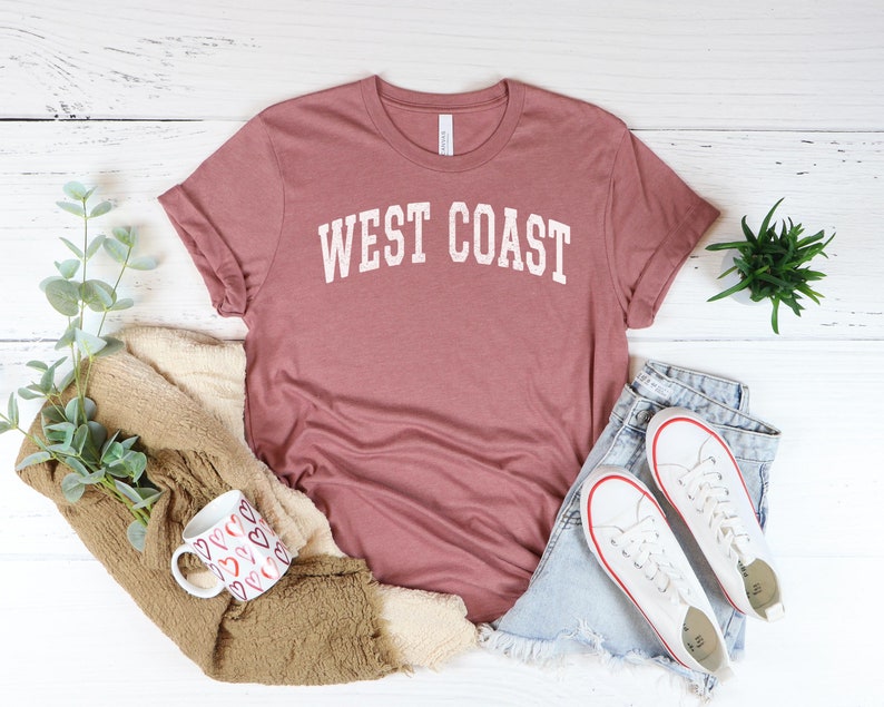 May include: A mauve colored t-shirt with the text "WEST COAST" printed in white on the front. The t-shirt is laid flat on a white wooden surface with a pair of white sneakers, a mug, and a plant.