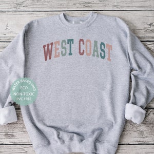 Puede incluir: Sudadera gris con el texto "WEST COAST" en colores arco&iacute;ris. La sudadera est&aacute; hecha con tintas a base de agua que son ecol&oacute;gicas, no t&oacute;xicas y libres de PVC.