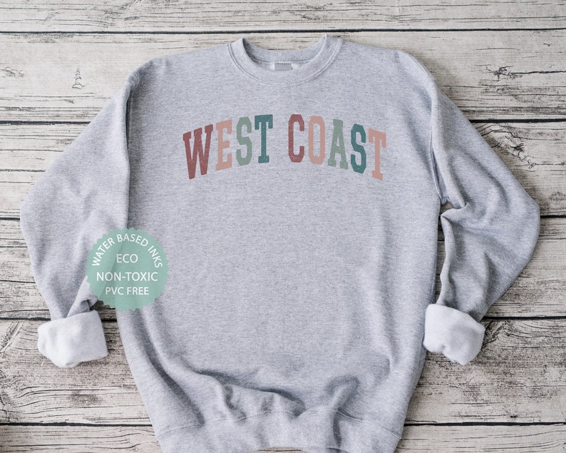 Puede incluir: Sudadera gris con el texto "WEST COAST" en colores arco&iacute;ris. La sudadera est&aacute; hecha con tintas a base de agua que son ecol&oacute;gicas, no t&oacute;xicas y libres de PVC.